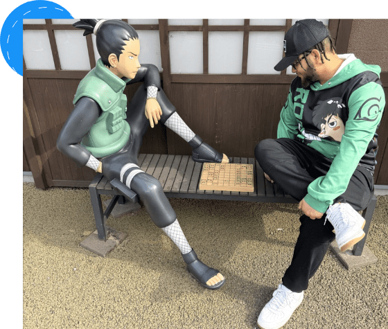 Two anime figures in green vests sitting and interacting by a wooden bench.